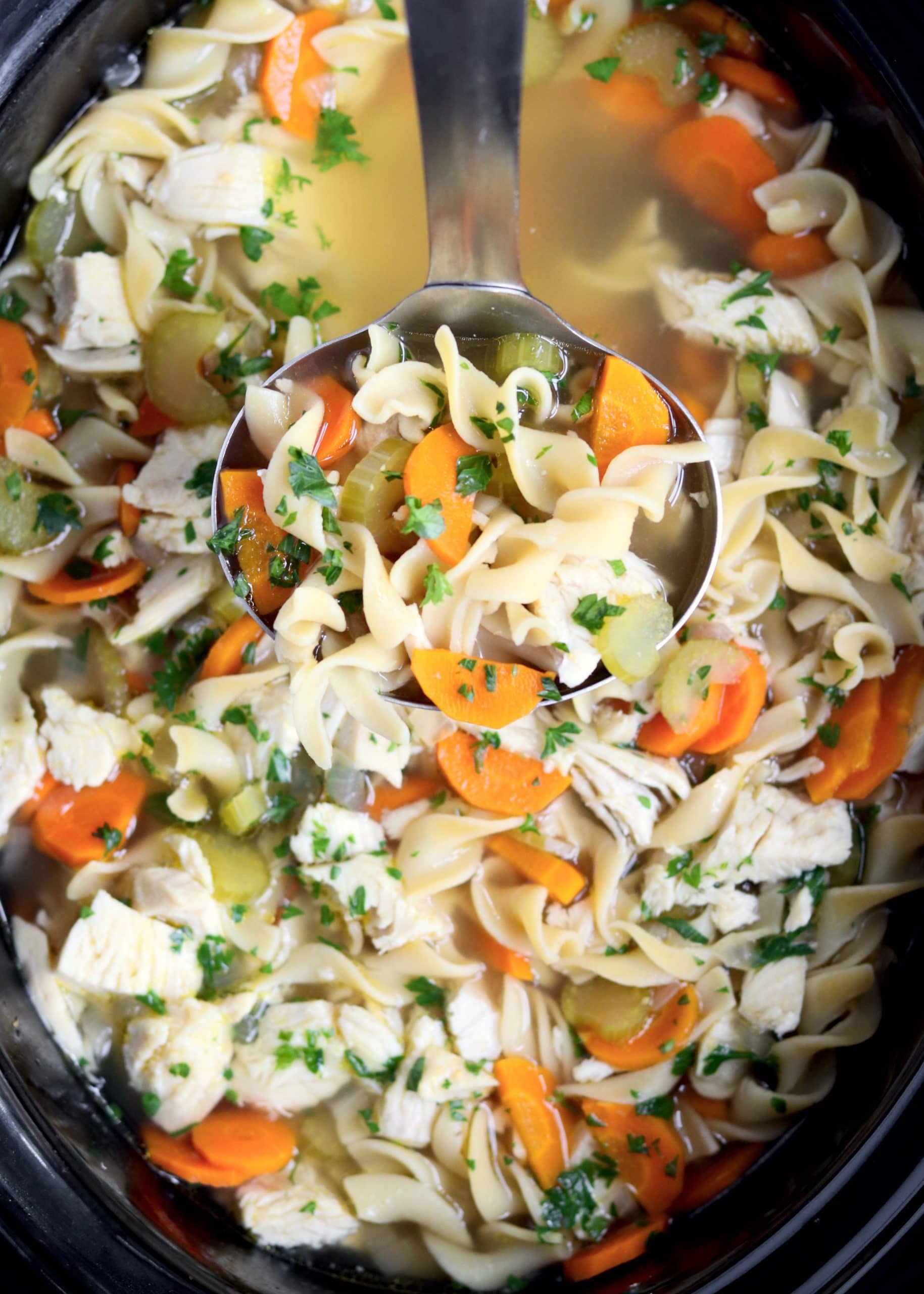 Ladle holding spoon of chicken noodle soup over a crockpot.