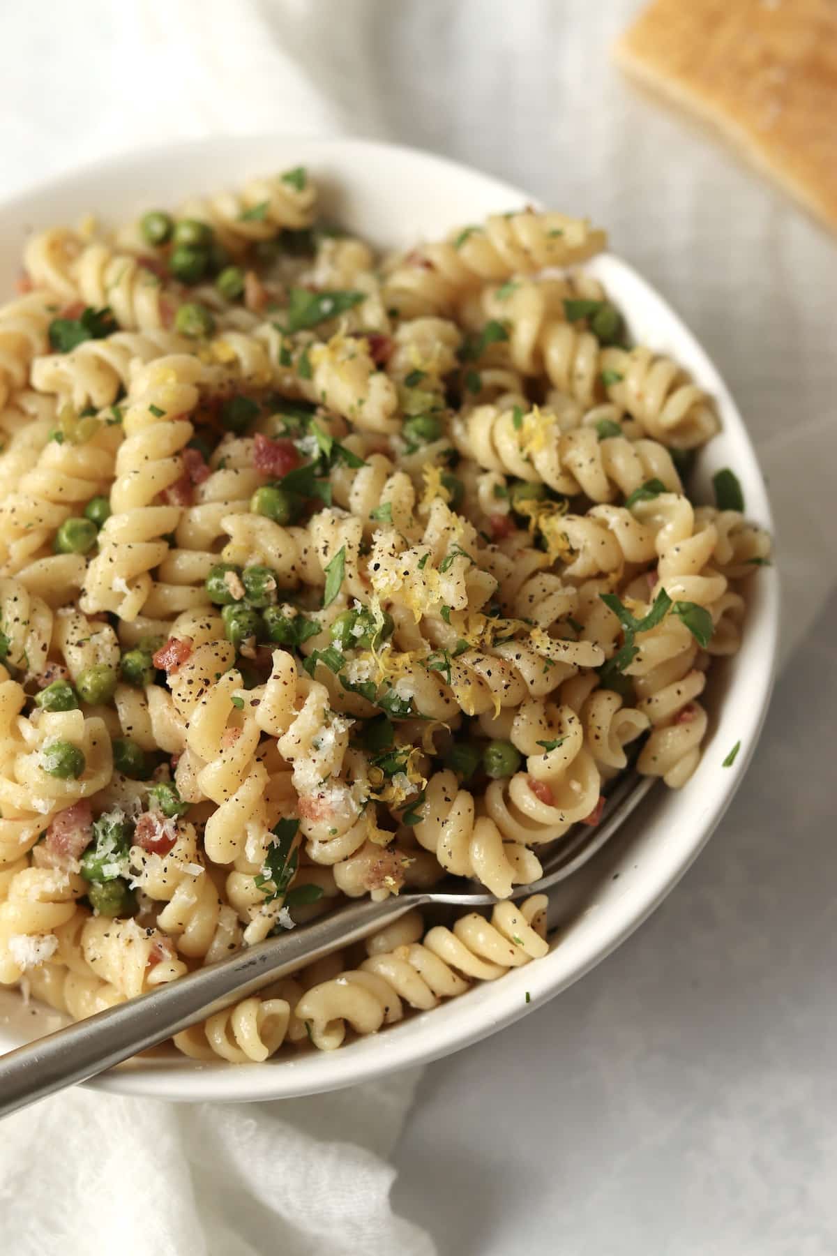 close up of rotini pasta with pancetta, peas, parsley, and lemon zest next to Parmesan Cheese