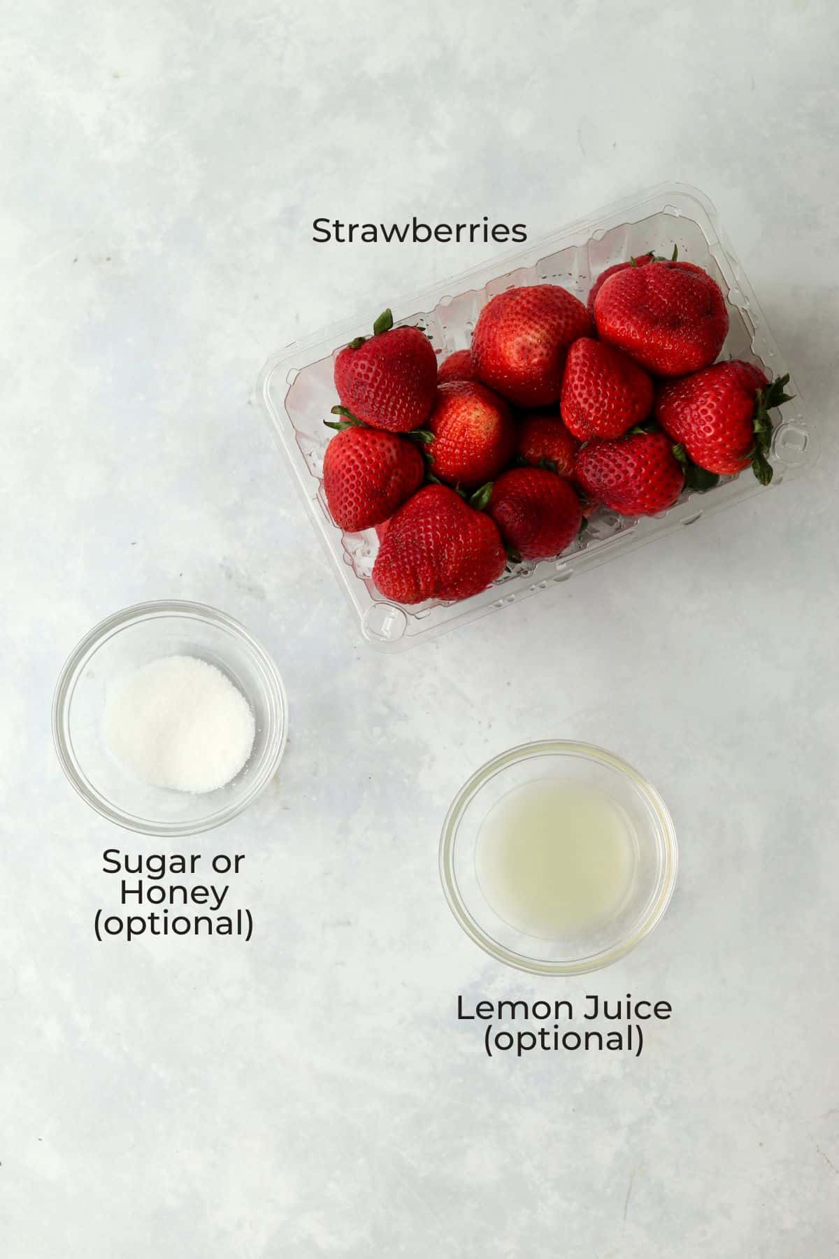 Fresh strawberries in their pint container, sugar in a small bowl, and lemon juice in a small bowl.
