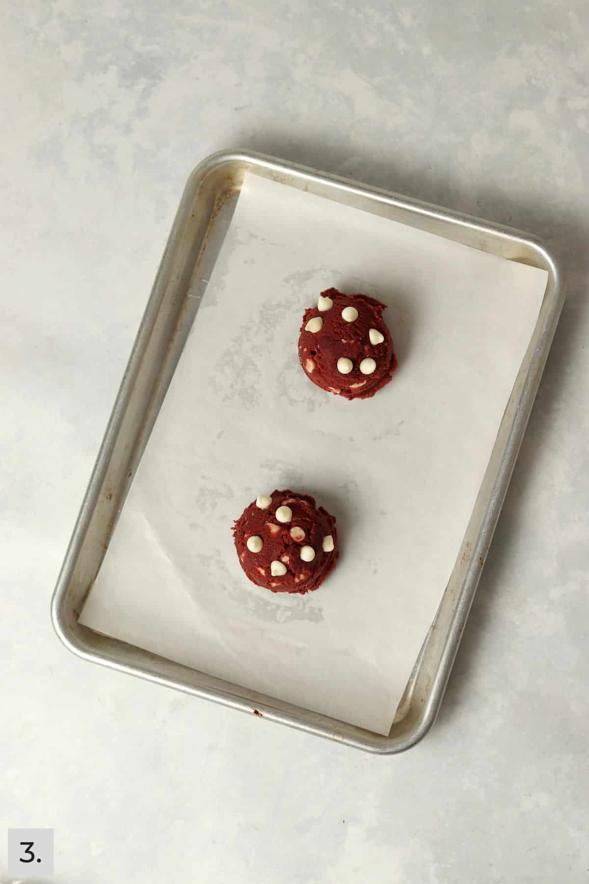 Two unbaked red velvet cookie dough balls on a baking sheet.