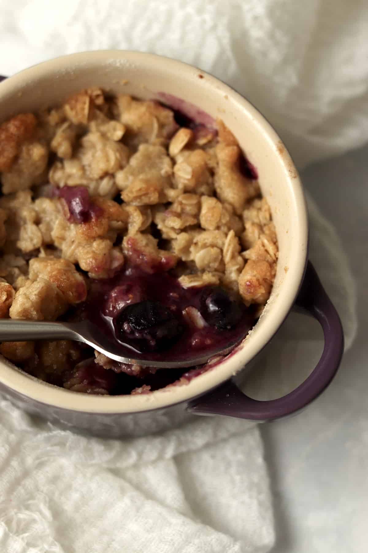 An individual blueberry crisp in a ramekin with a spoon.