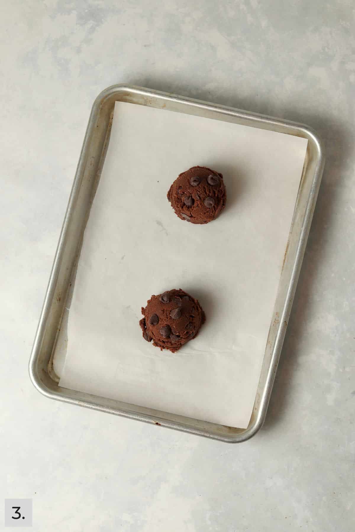 Two double chocolate cookie dough balls on a baking sheet.