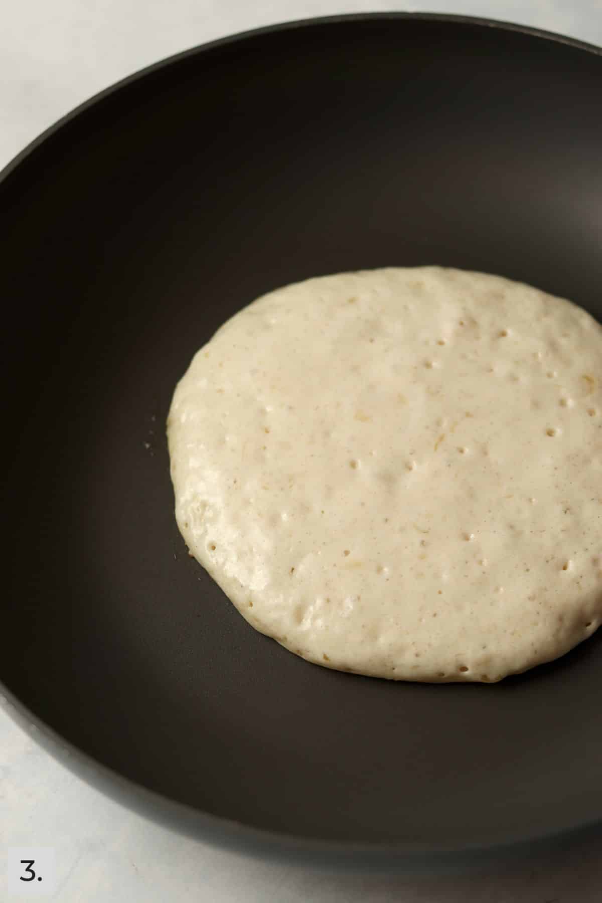 One pancake in a nonstick skillet before being flipped to show bubbles on top.