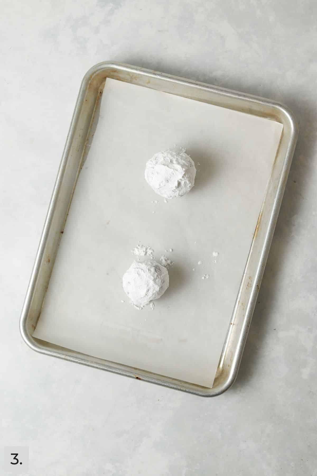 Two unbaked chocolate cookie dough balls rolled in powdered sugar on a parchment lined sheet.