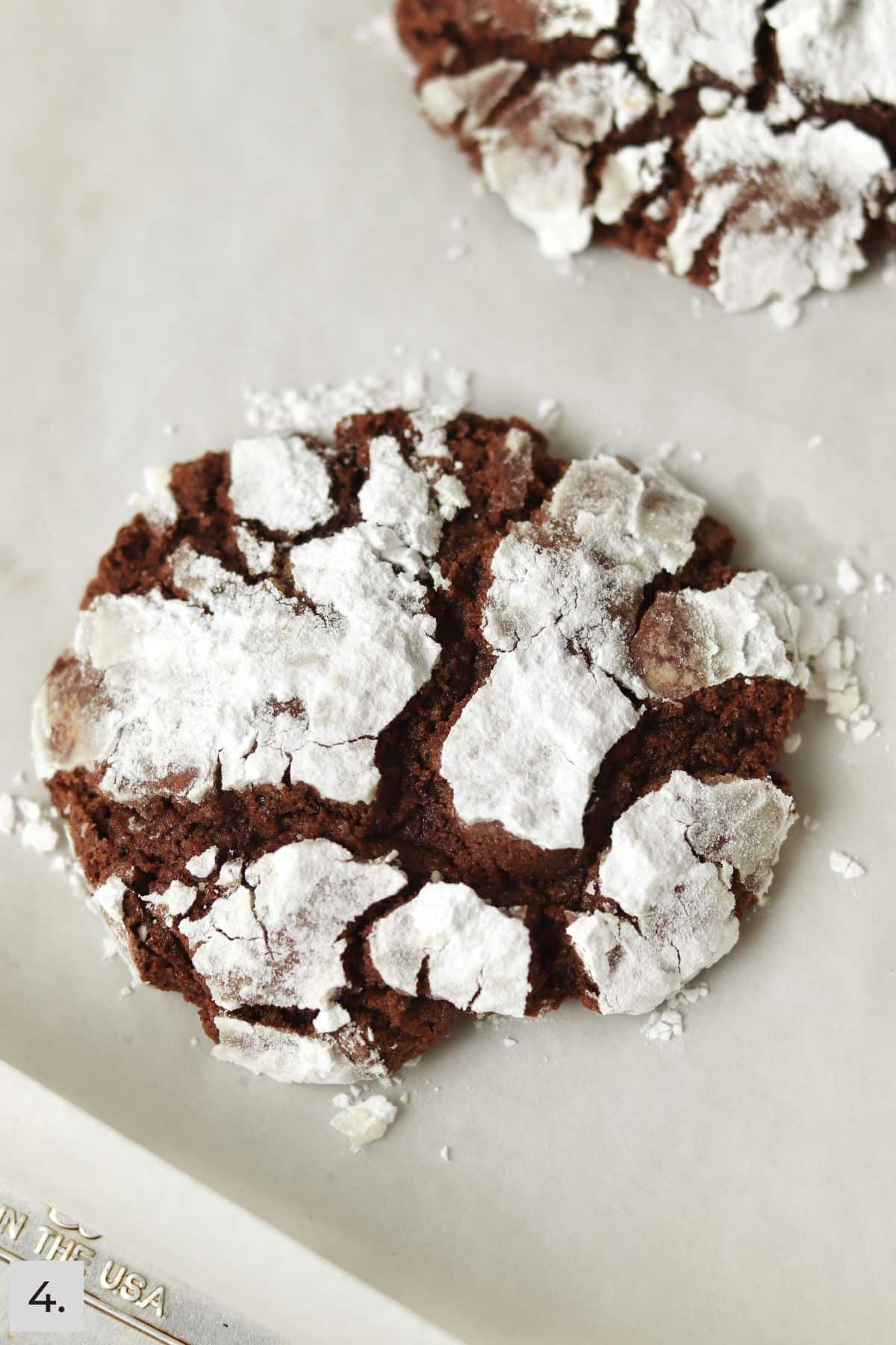 Two chocolate crinkle cookies on a parchment lined baking sheet.